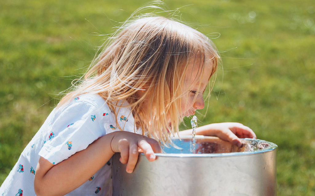 Kleines blondes Kind trinkt an einem Trinkbrunnen im Grünen. Kleines blondes Kind trinkt an einem Trinkbrunnen auf dem Spielplatz an einem heißen Sommertag.