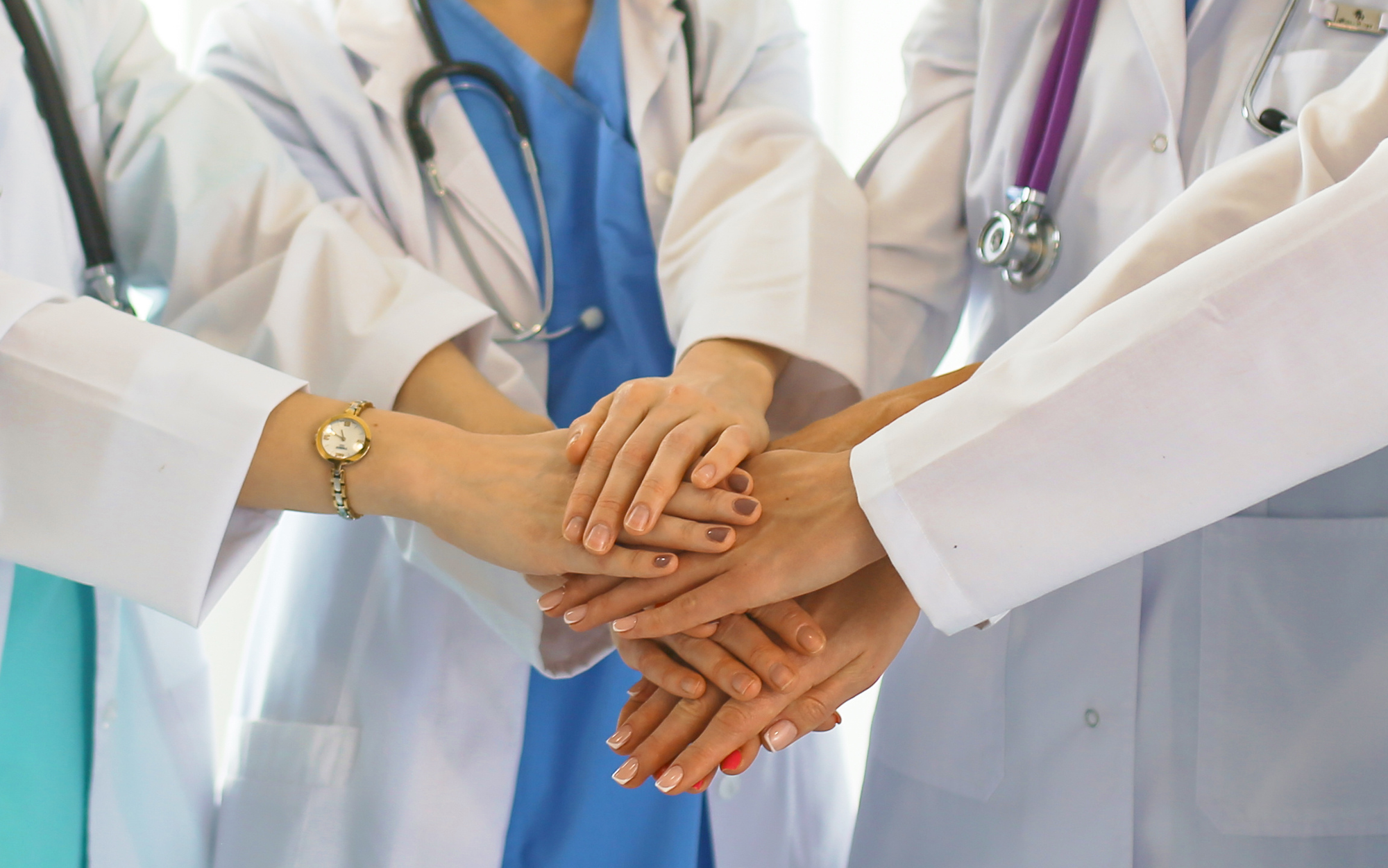 Mehrere Ärztinnen und Ärzte in weißen Kitteln legen ihre Hände übereinander als Zeichen von Teamarbeit und Zusammenhalt.