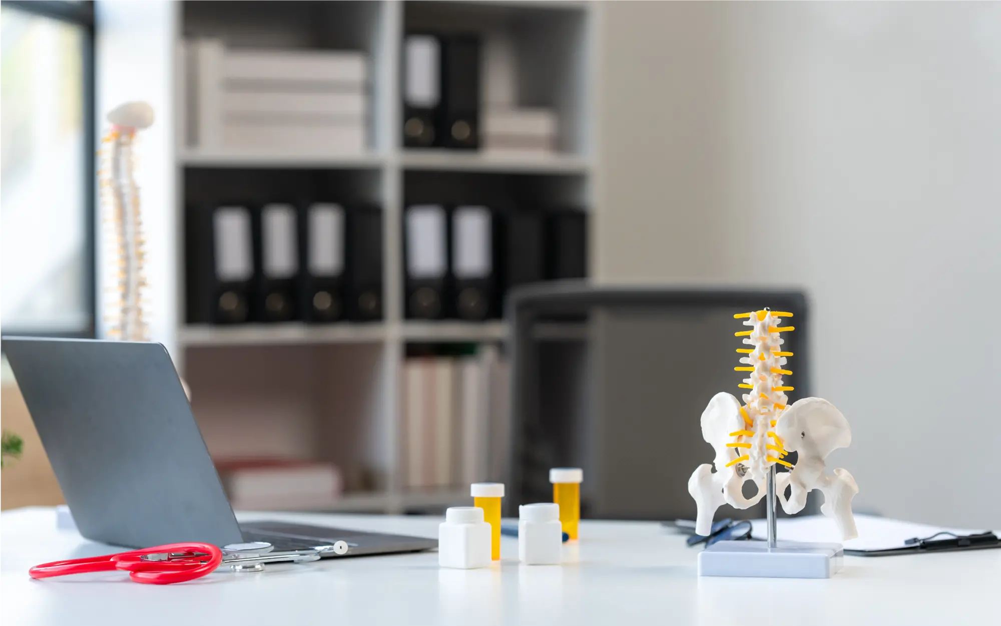 Schreibtisch in einer Arztpraxis mit einem anatomischen Modell der Wirbelsäule, Computer und typischer Büroausstattung.