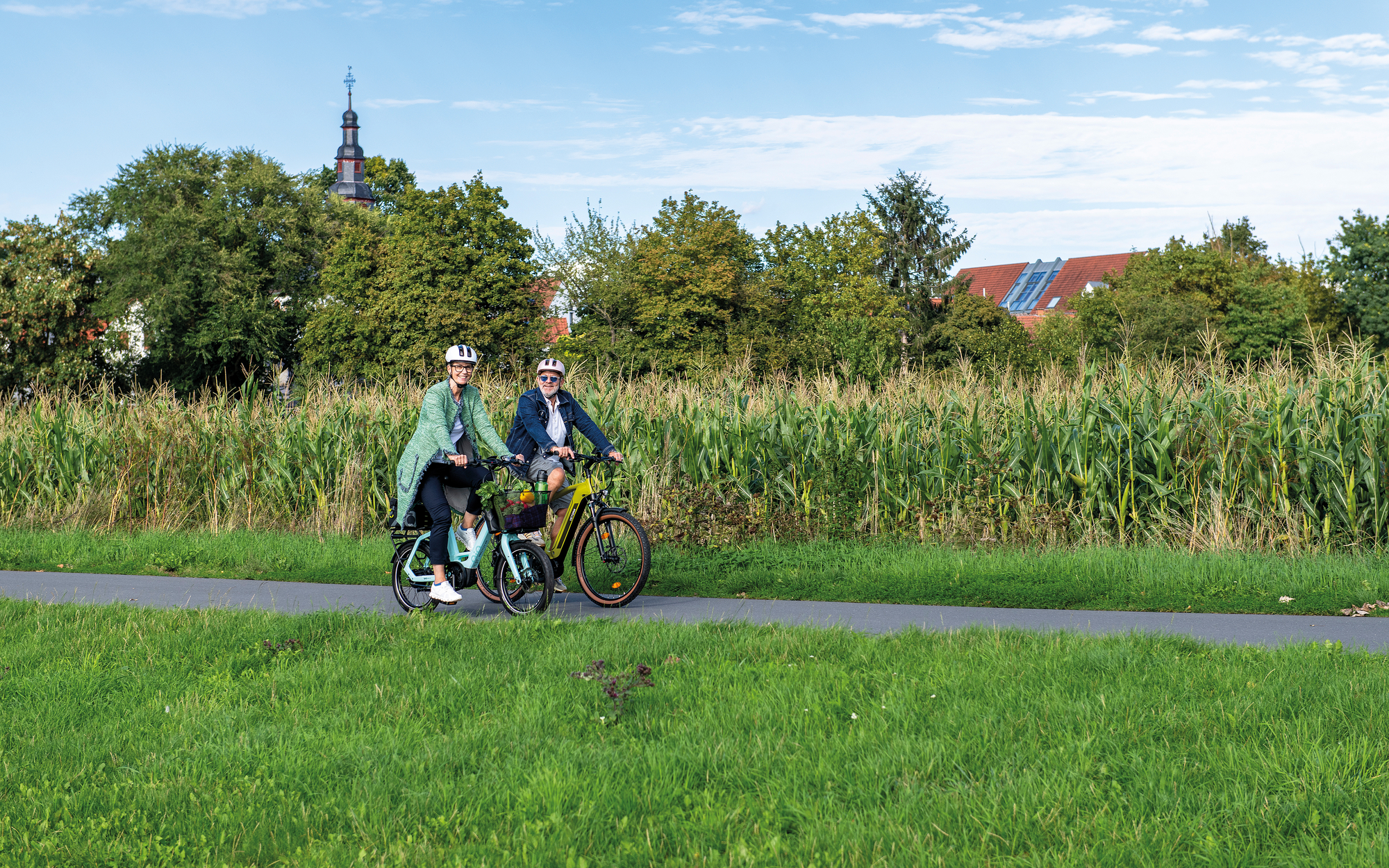 Zwei ältere Personen fahren mit ihren Fahrrädern und Einkäufen auf einem Weg durch ein weites, grünes Feld. Die Szene wirkt ruhig und naturverbunden.