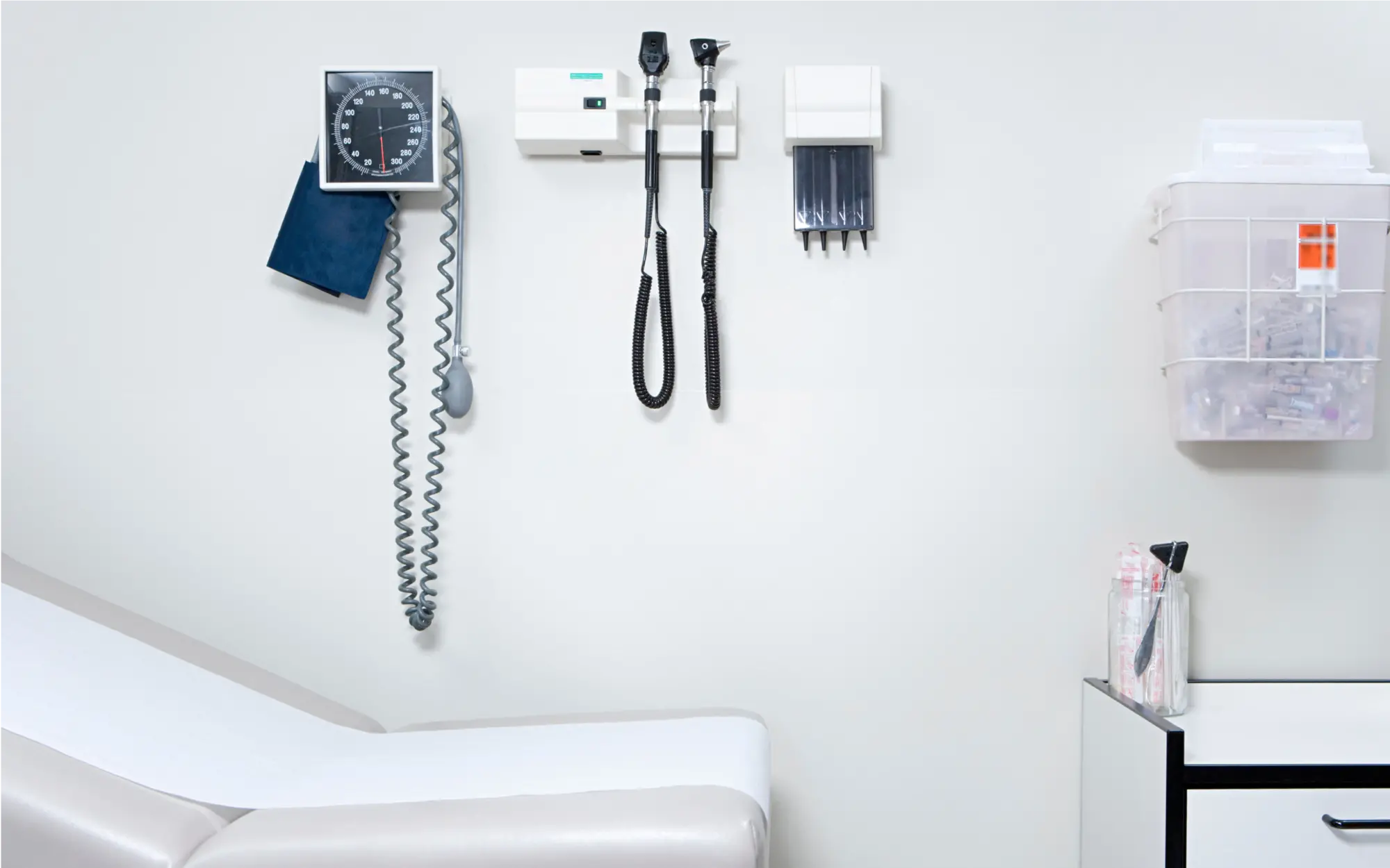 Medizinische Geräte und Ausstattung in einem Behandlungszimmer einer Arztpraxis, darunter Untersuchungsliege, Stethoskop, Blutdruckmessgerät und weitere typische Praxisgeräte.